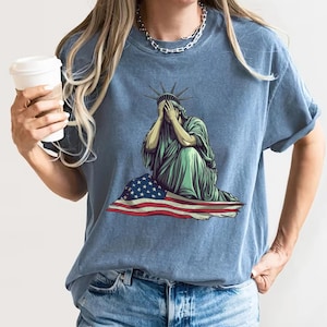 Emotional graphic of the Statue of Liberty sitting with her face in her hands wrapped in a tattered American flag symbolizing sorrow or concern for the nation perfect for political statement shirts freedom of speech tees protest art apparel or USA patriotism designs that express American values or disappointment in the state of the country