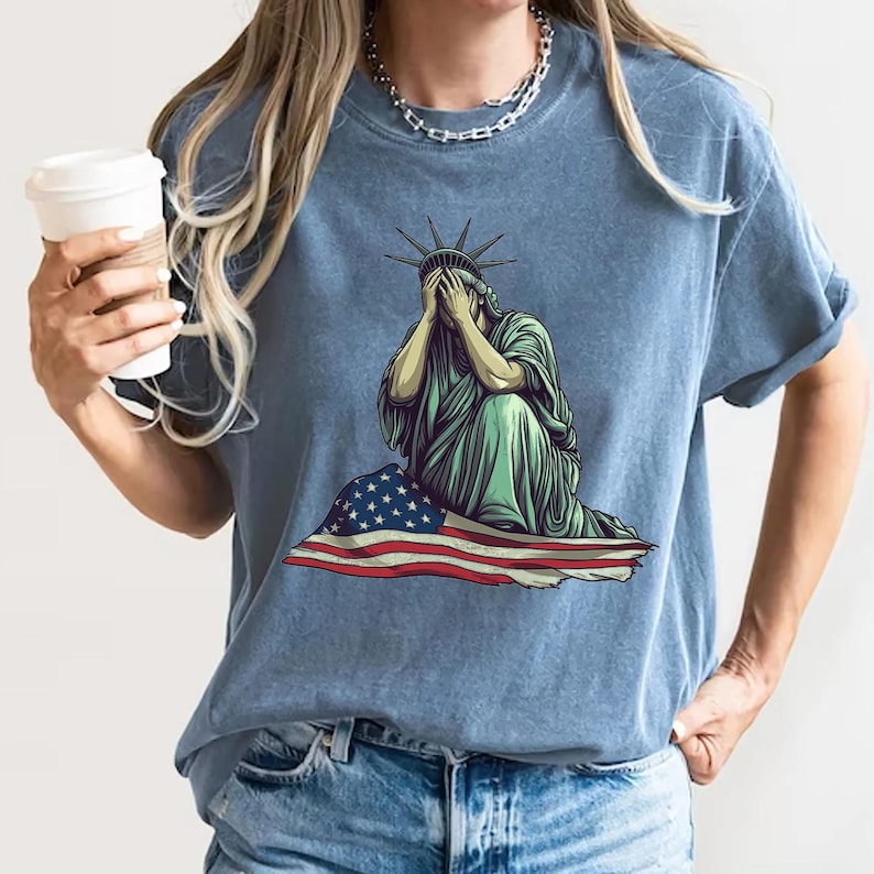 Emotional graphic of the Statue of Liberty sitting with her face in her hands wrapped in a tattered American flag symbolizing sorrow or concern for the nation perfect for political statement shirts freedom of speech tees protest art apparel or USA patriotism designs that express American values or disappointment in the state of the country