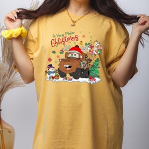 Festive holiday T-shirt featuring Disney’s Mater from Cars, wrapped in Christmas lights, with snowman and presents, perfect for Disney lovers.