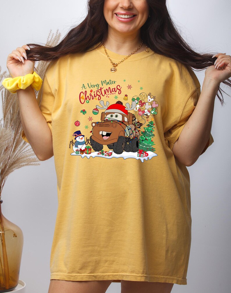 Festive holiday T-shirt featuring Disney’s Mater from Cars, wrapped in Christmas lights, with snowman and presents, perfect for Disney lovers.