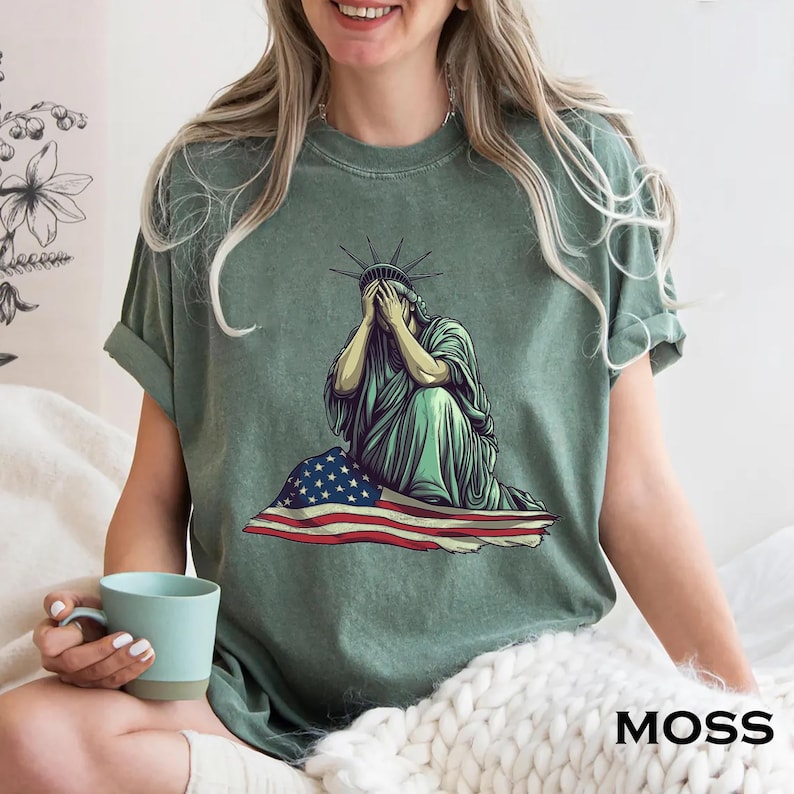 Emotional graphic of the Statue of Liberty sitting with her face in her hands wrapped in a tattered American flag symbolizing sorrow or concern for the nation perfect for political statement shirts freedom of speech tees protest art apparel or USA patriotism designs that express American values or disappointment in the state of the country