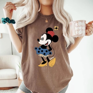 May include: A brown t-shirt featuring a graphic of Minnie Mouse wearing a blue polka dot skirt and yellow shoes. The graphic includes the text "Disney".