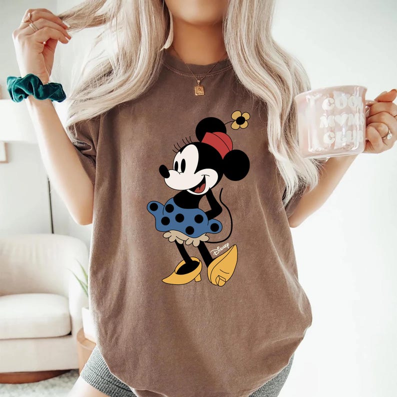 May include: A brown t-shirt featuring a graphic of Minnie Mouse wearing a blue polka dot skirt and yellow shoes. The graphic includes the text "Disney".