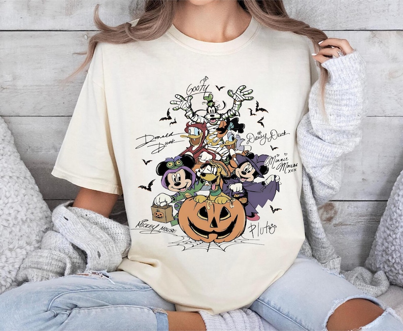 Retro-style Halloween design featuring Mickey Mouse, Minnie, Goofy, Donald, Daisy, and Pluto in costumes around a large jack-o'-lantern with bats and spiderwebs. Perfect for Disney Halloween party trips and fall-themed outfits.