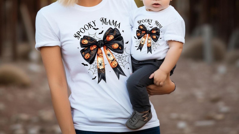 Op de afbeelding: Een wit T-shirt met een zwarte strik en oranje pompoenen, met de tekst "Spooky Mama" erop gedrukt. Een wit T-shirt met een zwarte strik en oranje pompoenen, met de tekst "Spooky Mini" erop gedrukt.