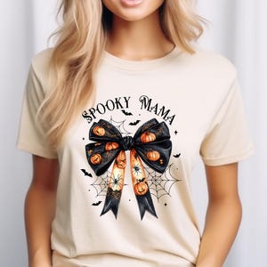 Op de afbeelding: Een beige T-shirt met een zwarte strik met oranje pompoenen en spinnen erop. De tekst "Spooky Mama" staat boven de strik.