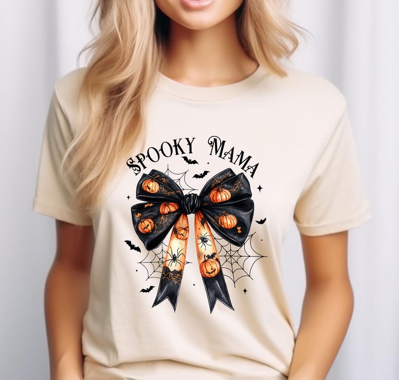 Op de afbeelding: Een beige T-shirt met een zwarte strik met oranje pompoenen en spinnen erop. De tekst "Spooky Mama" staat boven de strik.
