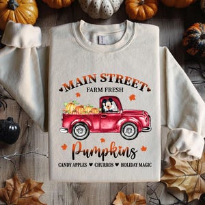 May include: A beige sweatshirt with a red truck carrying pumpkins and Mickey Mouse driving. The text on the sweatshirt says "Main Street Farm Fresh Pumpkins Candy Apples Churros Holiday Magic".