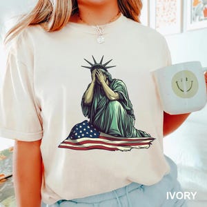 Emotional graphic of the Statue of Liberty sitting with her face in her hands wrapped in a tattered American flag symbolizing sorrow or concern for the nation perfect for political statement shirts freedom of speech tees protest art apparel or USA patriotism designs that express American values or disappointment in the state of the country