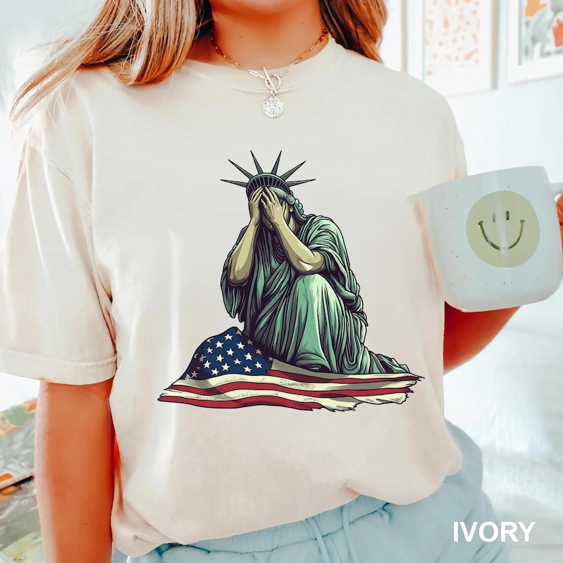 Emotional graphic of the Statue of Liberty sitting with her face in her hands wrapped in a tattered American flag symbolizing sorrow or concern for the nation perfect for political statement shirts freedom of speech tees protest art apparel or USA patriotism designs that express American values or disappointment in the state of the country