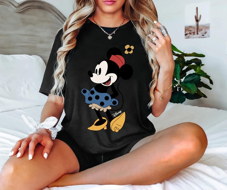 May include: Black t-shirt featuring a graphic of Minnie Mouse wearing a blue polka dot skirt with the word "Disney" printed on the bottom.
