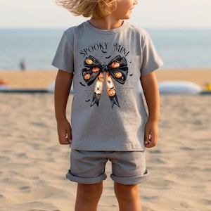 Op de afbeelding: Een grijs T-shirt met een zwart strikdesign met oranje en zwarte pompoenen en de tekst "Spooky Mini".