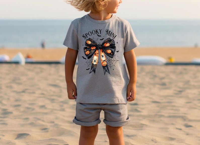 Op de afbeelding: Een grijs T-shirt met een zwart strikdesign met oranje en zwarte pompoenen en de tekst "Spooky Mini".