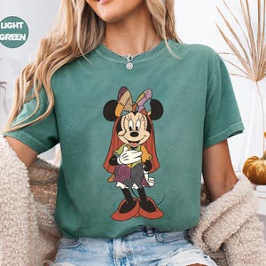 May include: Light green t-shirt featuring a Minnie Mouse graphic in a Halloween costume. The cartoon character has a patchwork bow, dress, and cape. The shirt has a round neckline and short sleeves. The words "LIGHT GREEN" are in a circle in the upper left corner.