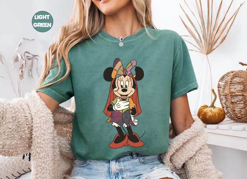 May include: Light green t-shirt featuring a Minnie Mouse graphic in a Halloween costume. The cartoon character has a patchwork bow, dress, and cape. The shirt has a round neckline and short sleeves. The words "LIGHT GREEN" are in a circle in the upper left corner.