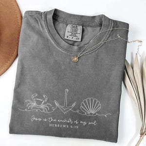 Puede incluir: Camiseta gris Comfort Colors con un dibujo lineal blanco de un cangrejo, un ancla y una concha. El texto "Jesus is the anchor of my soul" y "HEBREWS 6:19" están impresos debajo. Un collar plateado con un colgante está sobre la camiseta.