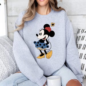May include: A grey sweatshirt featuring a cartoon image of Minnie Mouse wearing a red and white polka dot dress and a yellow flower in her hair. The image is centred on the sweatshirt.