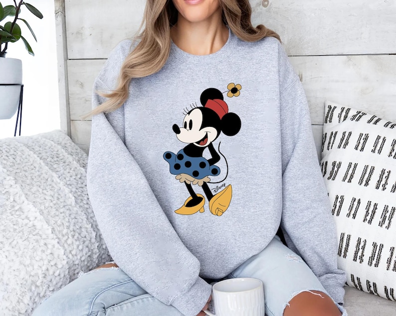May include: A grey sweatshirt featuring a cartoon image of Minnie Mouse wearing a red and white polka dot dress and a yellow flower in her hair. The image is centred on the sweatshirt.