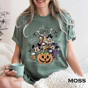 Retro-style Halloween design featuring Mickey Mouse, Minnie, Goofy, Donald, Daisy, and Pluto in costumes around a large jack-o'-lantern with bats and spiderwebs. Perfect for Disney Halloween party trips and fall-themed outfits.