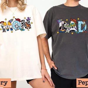 May include: Two t-shirts featuring a Toy Story theme. The white t-shirt says "MOM" with Toy Story characters. The grey t-shirt says "DAD" with Toy Story characters.