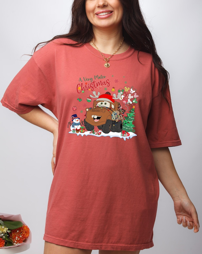 Disney Cars Christmas shirt featuring Mater in a festive holiday scene with lights, snowman, and gifts, perfect for Disney and Cars fans.