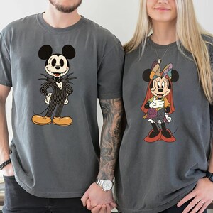 May include: Two gray t-shirts featuring Disney characters. One shirt displays Jack Skellington in a pinstripe suit, the other shows Minnie Mouse in a patchwork dress. Both shirts have short sleeves and a relaxed fit.