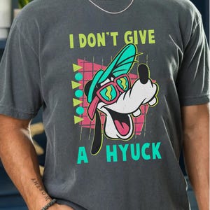 Retro 90s-style Disney shirt featuring Goofy wearing neon sunglasses and a sideways cap with bold text that says "I Don't Give A Hyuck." Colorful geometric background with a distressed vintage look. Perfect for fans of A Goofy Movie, Disney humor shirts, family vacations, or retro-themed Disney park outfits.