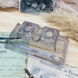 May include: A clear glass inkwell with two circular ink holders and a rectangular base. The inkwell sits on a wooden surface with vintage papers and a pen. A black and white photograph is in the background.