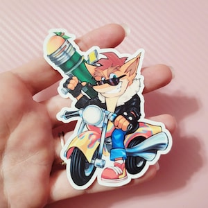 Könnte beinhalten: Ein Aufkleber mit einer Comicfigur auf einem Motorrad, die eine grüne Waffe hält. Die Figur hat orangefarbenes Fell, eine Sonnenbrille und eine schwarze Jacke. Das Motorrad ist gelb und rot mit Flammen.