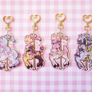 May include: Four colorful acrylic keychains featuring cartoon characters riding carousel animals. Each keychain has a gold heart-shaped clasp. The characters are a pink-haired girl riding a pink unicorn, a blonde girl riding a yellow and brown horse, a pink-haired girl riding a pink deer, and a dark-skinned girl riding a white and gold horse.