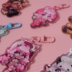 May include: A set of six colorful keychains featuring anime-style characters. Each keychain has a clear acrylic charm with a different character design and a metal ring for attaching to keys or bags.