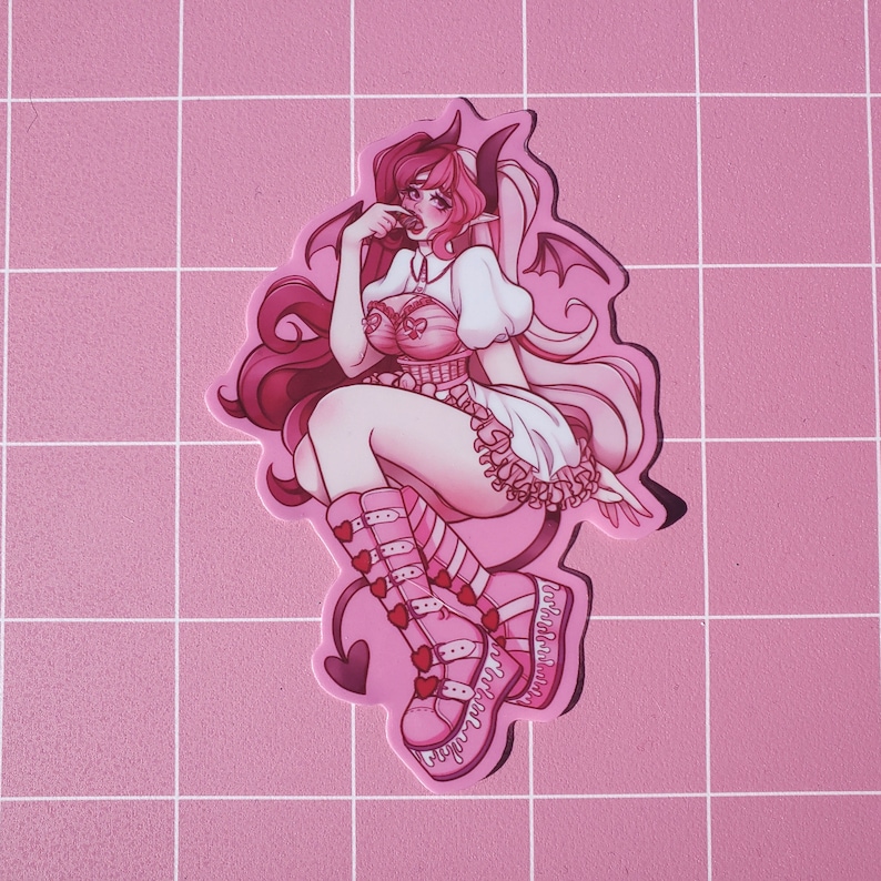 Puede incluir: Una pegatina de estilo anime rosa y blanco de un personaje femenino con cuernos, un vestido blanco y botas rosas. El personaje se muerde el dedo y tiene una expresi&oacute;n traviesa.