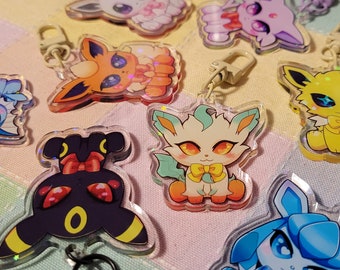 May include: A set of colorful anime-style character keychains featuring various Pokemon characters. The keychains are made of clear acrylic and feature a holographic effect. The characters include a yellow Eevee, a blue Espeon, a black Umbreon, a pink Sylveon, a red Flareon, and a white Glaceon.
