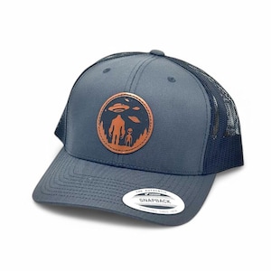 May include: A dark blue trucker hat with a brown leather patch featuring a silhouette of a person and an alien with a UFO above them. The text "The Authentic Snapback" is visible on the back of the hat.
