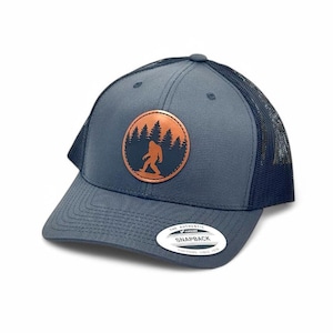 May include: A navy blue trucker hat with a brown leather patch featuring a silhouette of Bigfoot walking through a forest. The patch is circular and has a white outline. The hat has a black mesh back and the text "The Authentic Snapback" is printed on a white sticker on the back of the hat.
