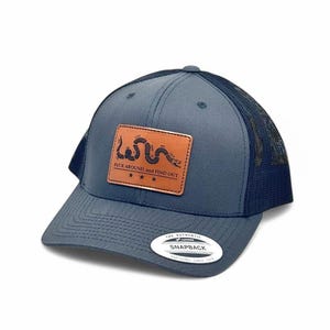 May include: A navy blue snapback trucker hat with a mesh back and a rectangular tan leather patch on the front. The patch features a black snake graphic, the text "FUCK AROUND, AND FIND OUT," and three stars. A white sticker is on the brim.