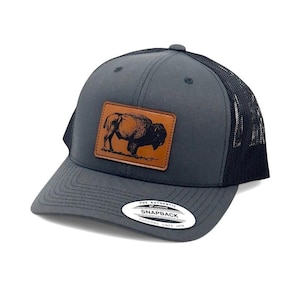 May include: A charcoal gray snapback cap featuring a rectangular brown leather patch on the front with a black engraved illustration of a standing bison. The cap has a black mesh back and a sticker on the brim that reads The Authentic Yupoong Snapback.