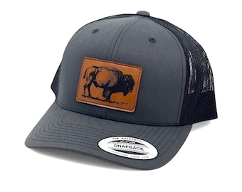 May include: A charcoal gray snapback cap featuring a rectangular brown leather patch on the front with a black engraved illustration of a standing bison. The cap has a black mesh back and a sticker on the brim that reads The Authentic Yupoong Snapback.