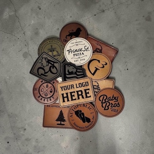 May include: A collection of brown and tan leather patches with various logos and text. Includes patches for "Prince St. Pizza", "Baby Bros Pizza & Wings", and "Your Logo Here". The patches are round, square, and rectangular.