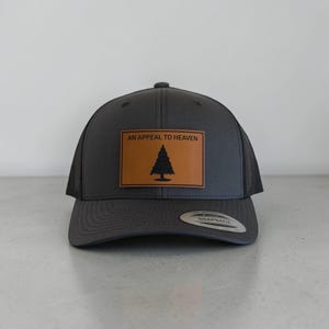 May include: A charcoal gray snapback cap featuring a rectangular tan leather patch on the front. The patch displays a black pine tree graphic and the text AN APPEAL TO HEAVEN. The cap has a mesh back and a circular silver sticker on the brim.
