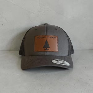 May include: A charcoal gray snapback cap featuring a rectangular brown leather patch on the front. The patch displays a black pine tree graphic and the text AN APPEAL TO HEAVEN. The hat has a mesh back and a sticker on the brim.