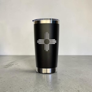 May include: Black stainless steel tumbler with a silver lid and base. The tumbler features a silver graphic of the Zia sun symbol, a Native American symbol. The tumbler is designed to keep beverages hot or cold.