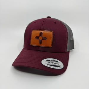 May include: A maroon and gray trucker hat with a brown leather patch featuring the symbol of the state of New Mexico. The hat has a mesh back and a curved brim. The hat has a sticker on the back that says "The Authentic Yupoong Snapback Yupoong Since 1974."