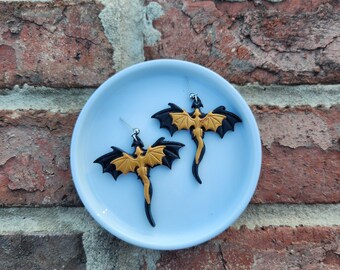 May include: A pair of gold and black dragon earrings on a blue plate. The dragons have black wings and gold bodies.
