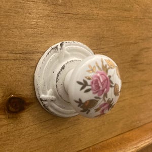 May include: White porcelain cabinet knob with a floral design featuring pink roses and gold accents. The knob has a decorative, ornate base.