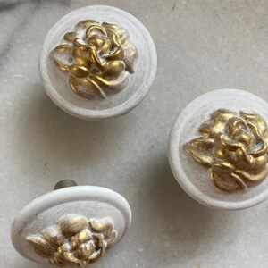 May include: Three white ceramic knobs with gold floral accents. The knobs are round and have a raised design.
