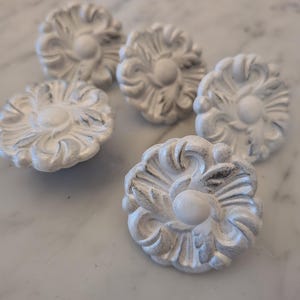 May include: A set of five circular cabinet knobs featuring a raised floral relief pattern. Each knob is finished in a matte white colour with subtle grey distressing in the recessed areas, highlighting the intricate carved petal details.