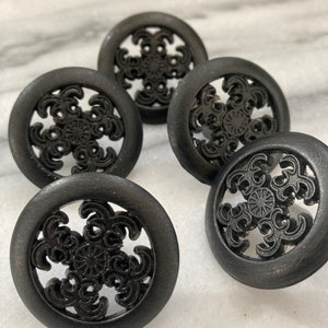 Peut inclure: Cinq boutons de meuble noirs décoratifs avec un design circulaire. Chaque bouton présente un motif ajouré avec un motif floral central. Les boutons sont faits d'un matériau sombre et mat et sont disposés sur une surface claire.