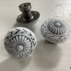 Peut inclure: Trois boutons de meuble décoratifs avec une finition blanche vieillie. Les boutons présentent un motif floral avec une base en métal foncé. Un bouton est démonté, montrant la quincaillerie métallique.