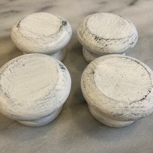 Peut inclure: Quatre boutons de meuble blancs vieillis, avec un dessus rond et une courte tige. Les boutons ont un aspect légèrement texturé avec des nuances grises visibles, suggérant un style rustique ou de ferme. Ils sont disposés en carré.